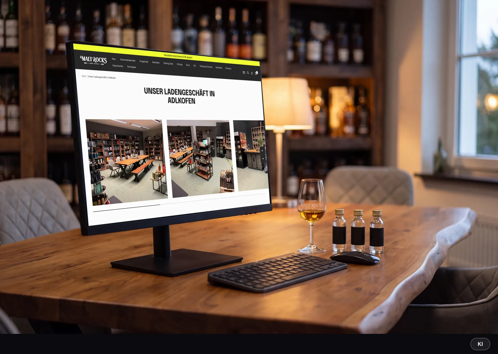 KI-generiertes Bild: Monitor auf einem Schreibtisch zeigt die Seite zum Malt-Rocks-Ladengeschäft in Adlkofen mit Fotos des Ladens, davor Tastatur, Nosing-Glas und Sample-Fläschchen