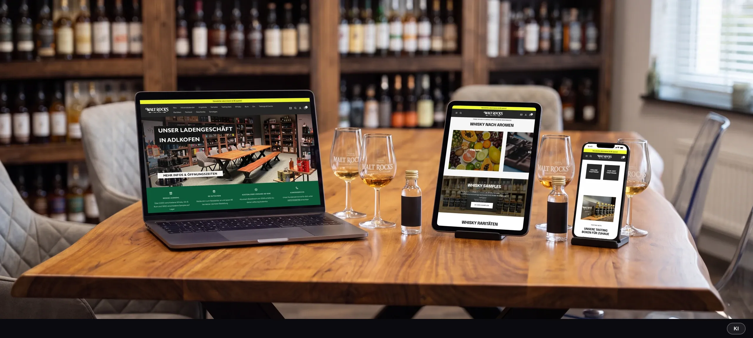 KI-generiertes Bild: Laptop, Tablet und Smartphone vor Spirituosenregalen zeigen die Malt-Rocks-Startseite, die Whisky-Auswahl nach Aromen und die Tastings